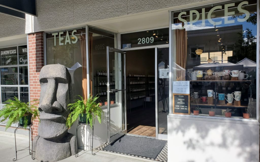 a photo of the outside a tea shop called Chai Cupboard, with a large replica Moai on the sidewalk in front of it, flanked by two ferns